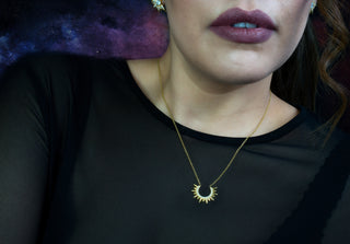 Close-up of a woman wearing a gold necklace with a sun pendant against a dark background.